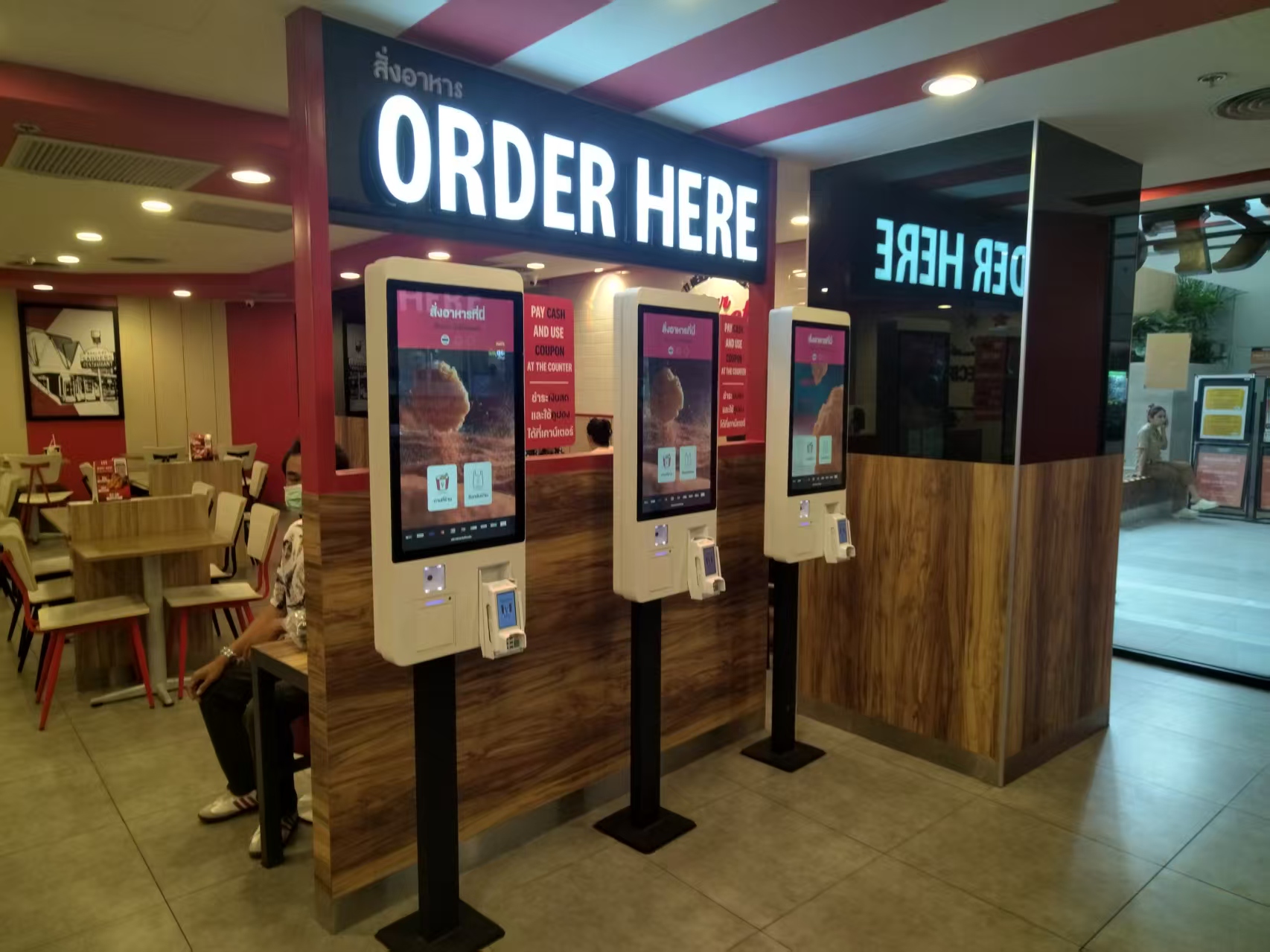 Three touchscreen self-ordering terminals beneath an Order Here sign in a restaurant